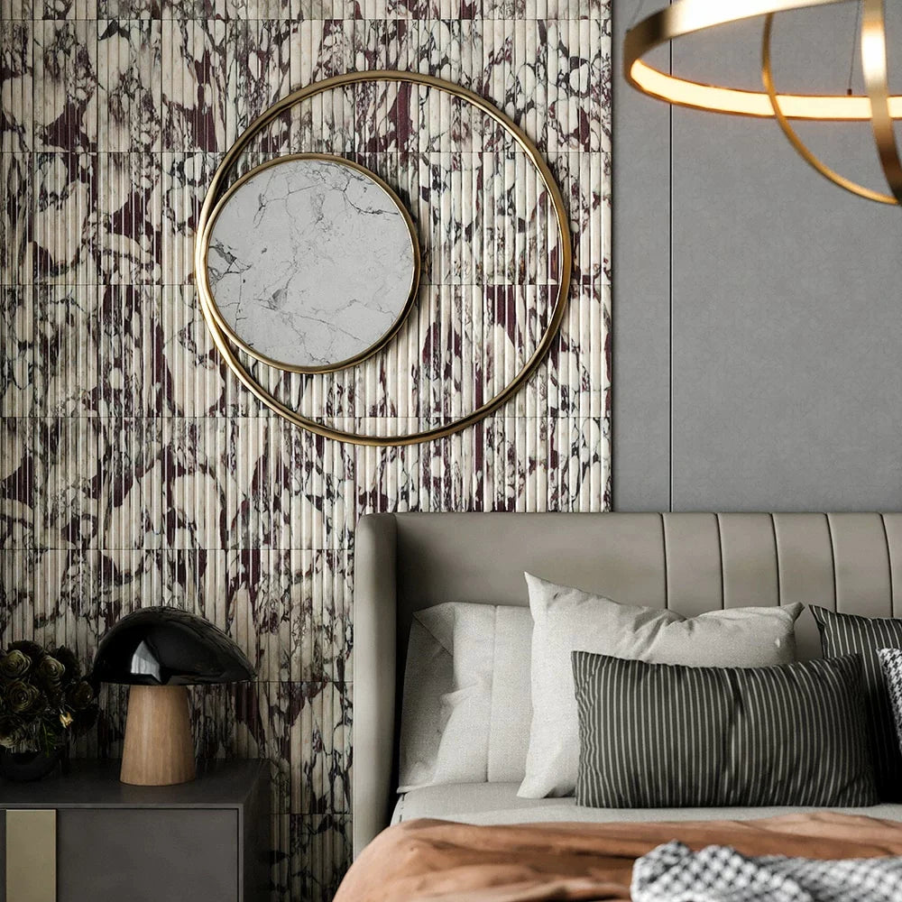 Modern bedroom with fluted wall tiles in white, gray, and burgundy, gray upholstered bed, round mirror with gold accents, and black bedside lamp.
