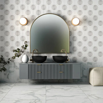 Modern bathroom with patterned wall tiles, large arched mirror, dual black sinks, gold faucets, gray floating vanity, marble floor tiles, round wall sconces.