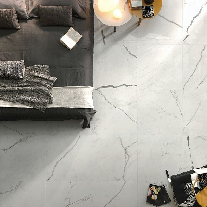 Modern bedroom with large white marble tiles, dark gray bedding, and sleek aesthetics. Includes round side table with lamp and books.