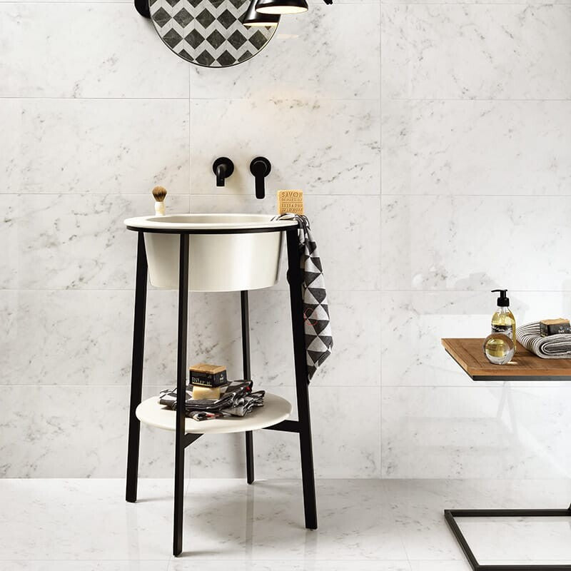 Modern bathroom with white marble tiles, white basin on black legs, round mirror, wooden side table with toiletries, and black wall-mounted faucet.