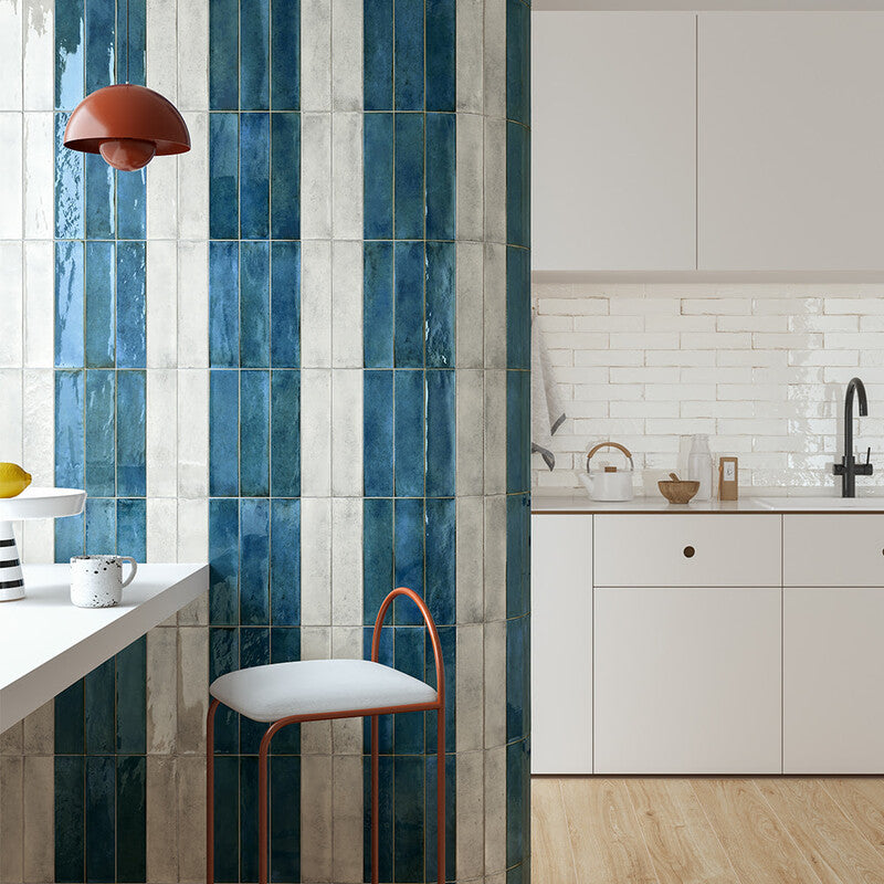 Modern kitchen with white countertop, cabinets, red accents. Striped wall with blue/white tiles, sleek bar stool, red pendant light, black faucet.