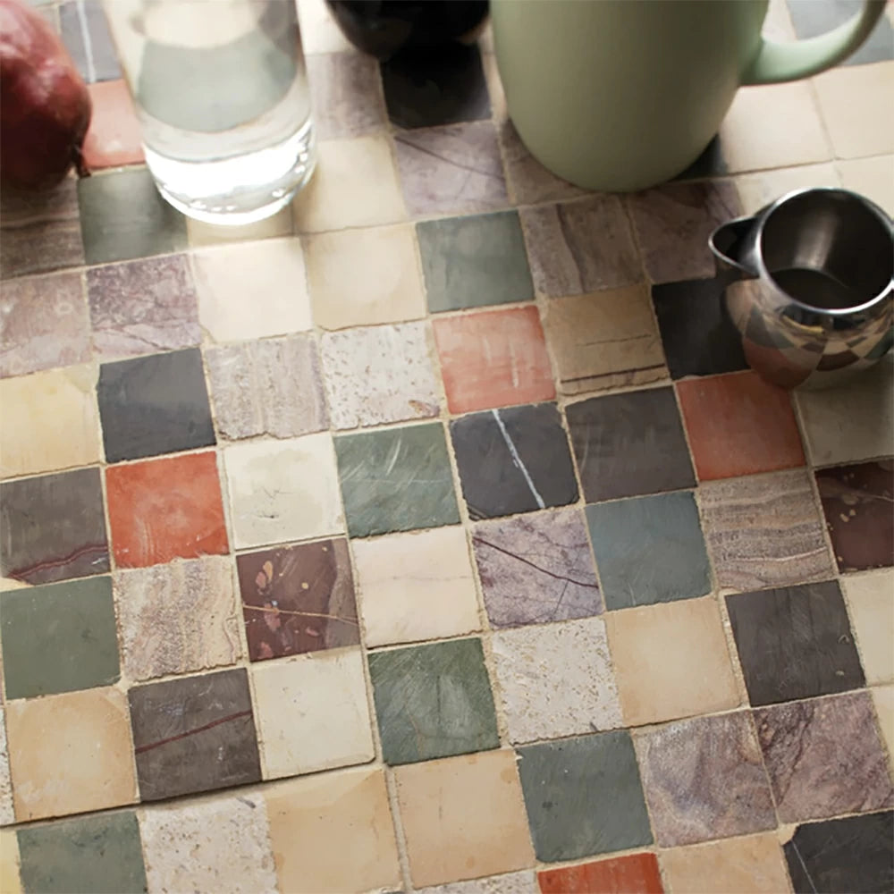 Close-up of mosaic tile surface with small square tiles in beige, red, green, and gray. Also visible: glass of water, green mug, metal pitcher, red onion.