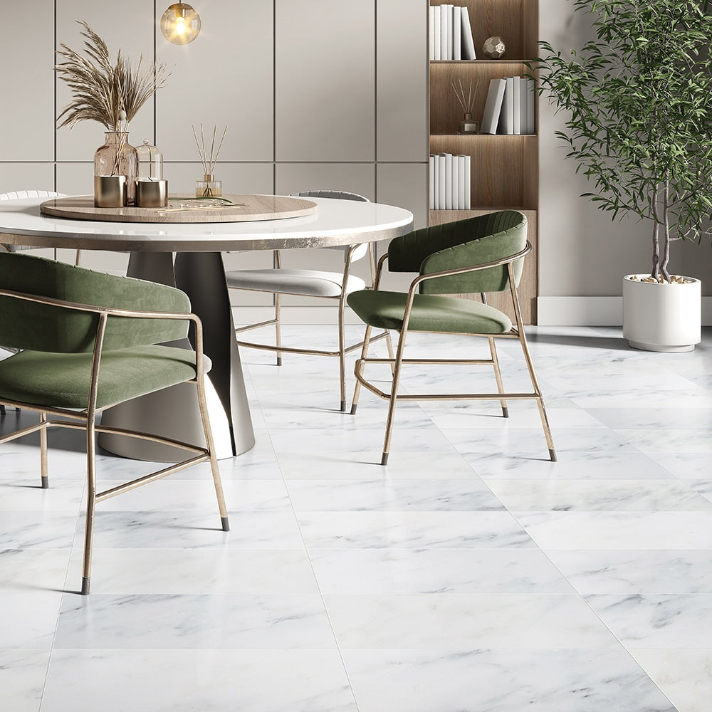 Modern dining area with white marble tile flooring, green velvet chairs, round white table, bookshelf with decor, and a potted plant.