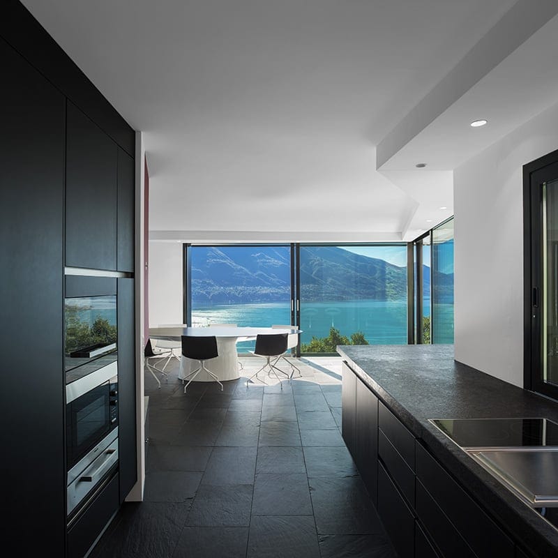 Modern kitchen with black slate tile flooring, dark cabinetry, and large windows with scenic lake and mountain views.