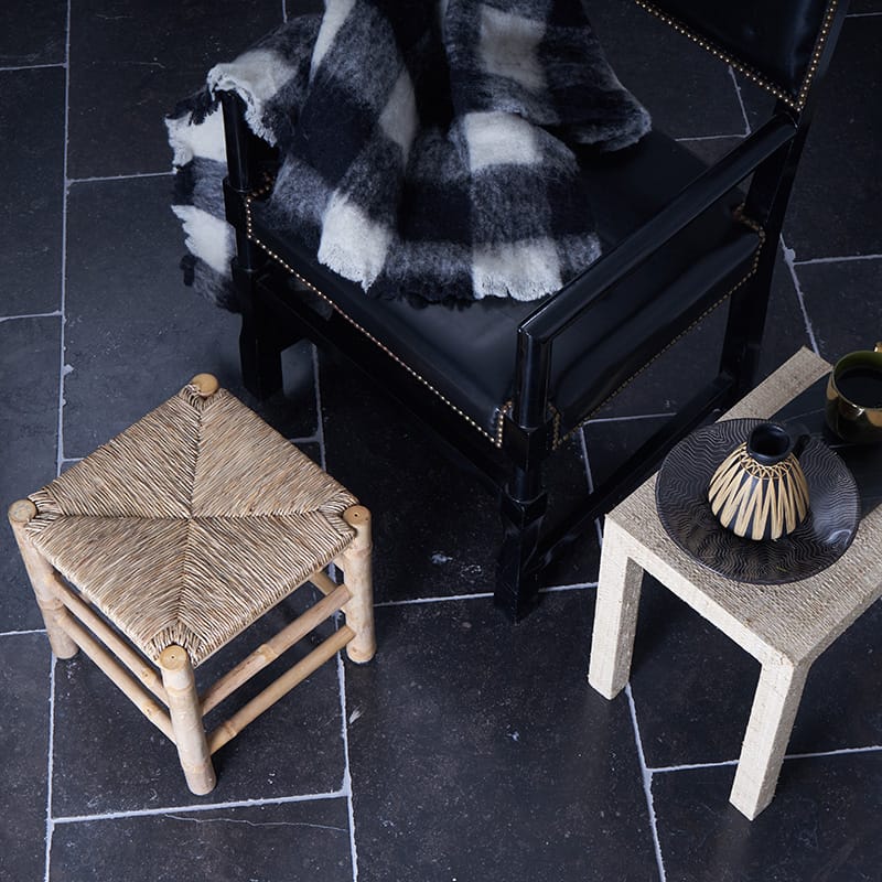 Black leather chair with brass nailhead trim, checkered blanket, stone table with black and white vase, wooden stool on grey tile floor.