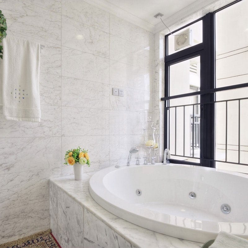 Luxurious bathroom with white Carrara marble tiles, large round bathtub, chrome fixtures, black-framed window, towel, vase with yellow flowers.