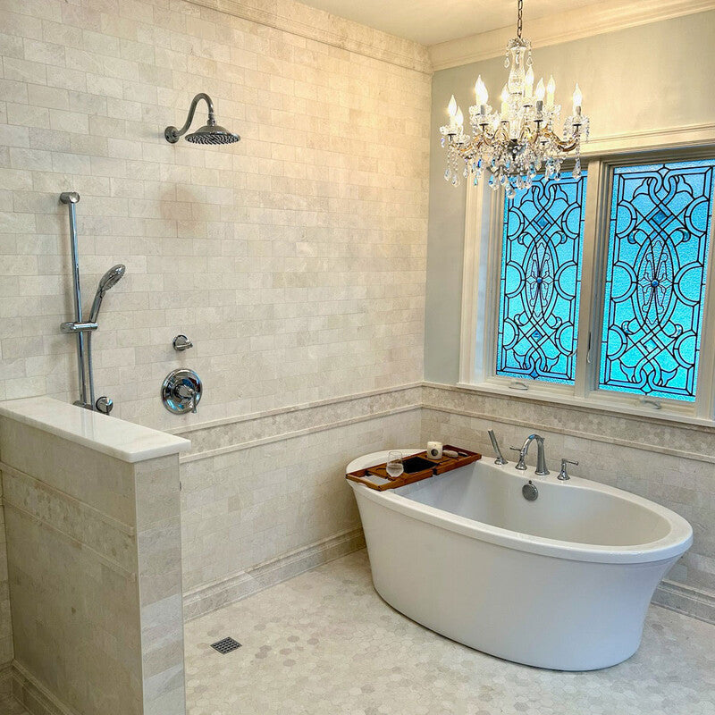 Luxurious bathroom with white bathtub, wooden caddy, chandelier, chrome fixtures, beige marble tiles, and stained glass window for natural light.