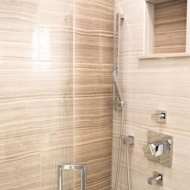 Modern bathroom with beige marble-look tiles, built-in shelf, and shiny chrome fixtures including showerhead and faucet.