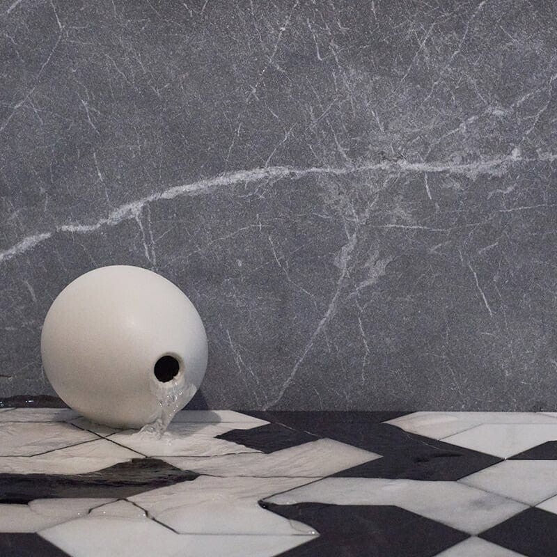 White vase pours water on geometric tiles in black, white, and grey with various finishes. Background is grey stone wall with white veining.