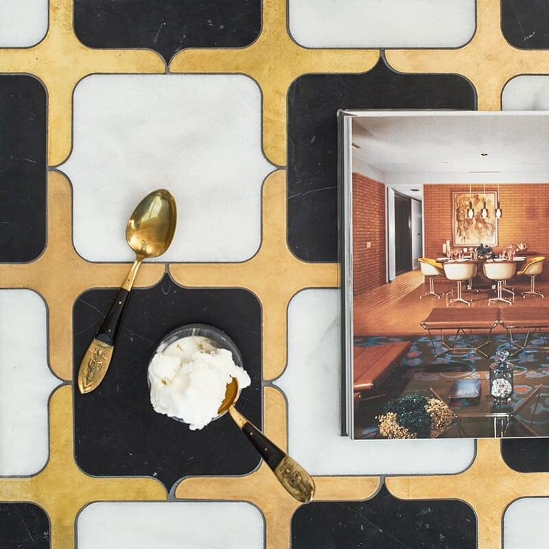 Close-up of black and white marble tile with gold geometric patterns, gold spoon and white cream on one side, magazine with modern dining room on the other.