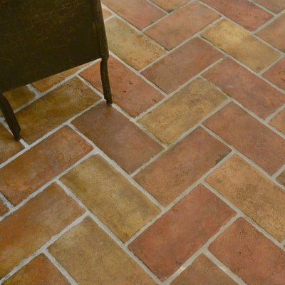 Reclaimed terracotta tiles in herringbone pattern with red, brown, yellow shades, bordered by dark wood chair leg.